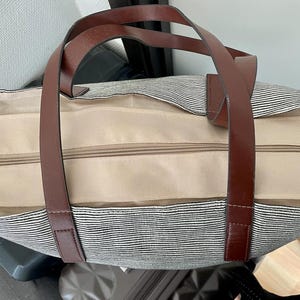 以下が含まれることがあります： 茶色の革のハンドルとジッパーの留め具が付いた、ストライプのキャンバス地のトートバッグ。丈夫な素材で作られており、広々とした内装です。