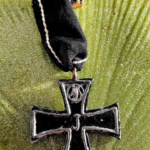 May include: A black Iron Cross medal with a silver border and a black ribbon. The medal has the year 1914 on the bottom and the letter "J" in the centre. The ribbon is black with white trim.