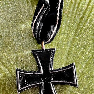 May include: A black Iron Cross medal with a white border and a black ribbon with white stripes. The medal is attached to the ribbon with a gold safety pin. The background is a textured green surface.