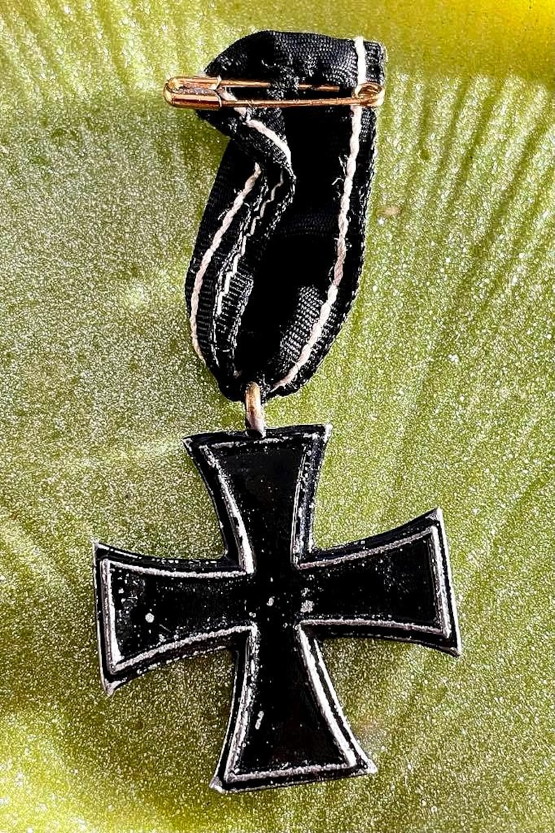 May include: A black Iron Cross medal with a white border and a black ribbon with white stripes. The medal is attached to the ribbon with a gold safety pin. The background is a textured green surface.