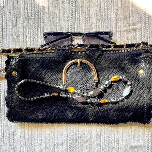 Peut inclure: Une pochette noire avec une boucle dor&eacute;e et une cha&icirc;ne, pr&eacute;sentant un motif de peau de serpent textur&eacute;. Des lunettes de soleil sont pos&eacute;es dessus, et un collier de perles avec des accents jaunes et argent&eacute;s est drap&eacute; sur le devant.