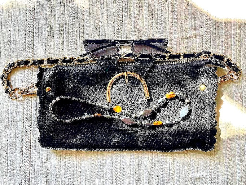 Peut inclure: Une pochette noire avec une boucle dor&eacute;e et une cha&icirc;ne, pr&eacute;sentant un motif de peau de serpent textur&eacute;. Des lunettes de soleil sont pos&eacute;es dessus, et un collier de perles avec des accents jaunes et argent&eacute;s est drap&eacute; sur le devant.