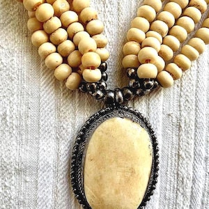 Puede incluir: Un collar de múltiples hilos hecho de cuentas de color crema con un gran colgante ovalado. El colgante está hecho de un material de color crema con un marco de metal plateado.