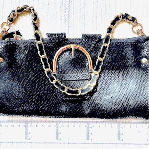 Könnte beinhalten: Schwarze Clutch mit goldfarbenem Kettenriemen und Schnallenverschluss. Die Tasche hat ein strukturiertes, schlangenartiges Muster und gewellte Kanten. Goldakzente sind am Reißverschluss und an den Seiten sichtbar. Ein Lineal liegt darunter.