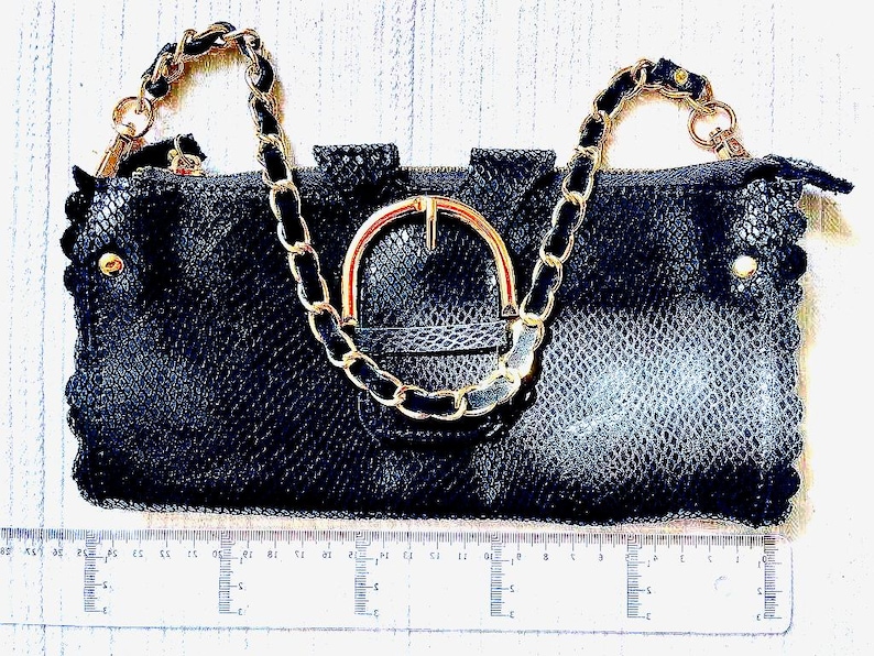 Peut inclure: Pochette noire avec une bandouli&egrave;re cha&icirc;ne et une boucle dor&eacute;e. Le sac pr&eacute;sente un motif textur&eacute; en forme de serpent et des bords festonn&eacute;s. Des accents dor&eacute;s sont visibles sur la fermeture &eacute;clair et les c&ocirc;t&eacute;s. Une r&egrave;gle est plac&eacute;e en dessous.