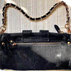 Peut inclure: Pochette noire avec une bandouli&egrave;re cha&icirc;ne dor&eacute;e. La pochette pr&eacute;sente un motif textur&eacute; en forme de peau de serpent et une fermeture &eacute;clair dor&eacute;e. La bandouli&egrave;re cha&icirc;ne est dor&eacute;e avec des accents de velours noir.