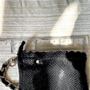 Peut inclure: Pochette noire avec une bandouli&egrave;re cha&icirc;ne. La pochette pr&eacute;sente un motif textur&eacute; en forme d'&eacute;cailles et un bord festonn&eacute;. Une r&egrave;gle est plac&eacute;e &agrave; c&ocirc;t&eacute; de la pochette, montrant une longueur d'environ 15 cm.