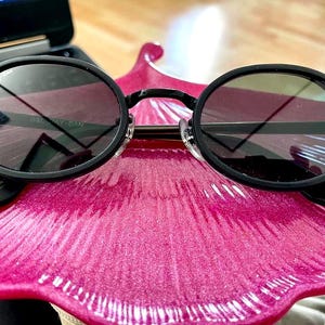 May include: Black round sunglasses with dark lenses. The sunglasses have a black frame and arms. The lenses reflect the surrounding environment. The sunglasses are resting on a pink, petal-shaped surface.