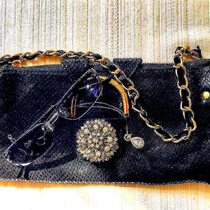 Peut inclure: Pochette noire avec une cha&icirc;ne dor&eacute;e, orn&eacute;e de lunettes, d'une broche en strass et d'un petit pendentif. La pochette pr&eacute;sente une surface textur&eacute;e et des d&eacute;tails dor&eacute;s.