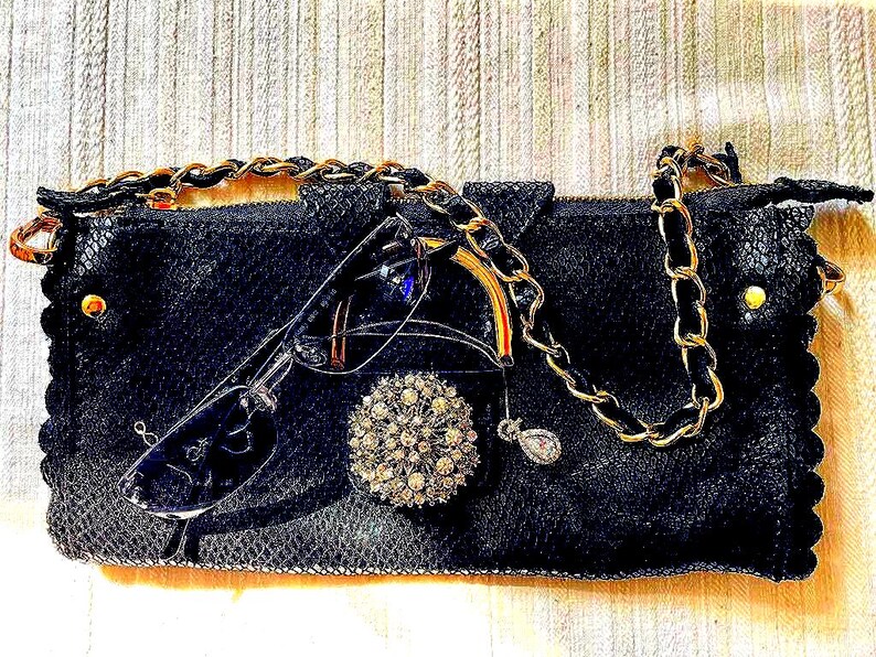 Peut inclure: Pochette noire avec une cha&icirc;ne dor&eacute;e, orn&eacute;e de lunettes, d'une broche en strass et d'un petit pendentif. La pochette pr&eacute;sente une surface textur&eacute;e et des d&eacute;tails dor&eacute;s.