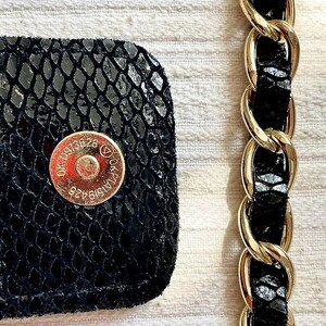 Peut inclure: Gros plan sur un d&eacute;tail de sac &agrave; main noir avec un motif textur&eacute; en peau de serpent. Une bandouli&egrave;re en cha&icirc;ne dor&eacute;e est visible &agrave; droite. Le sac comporte une fermeture magn&eacute;tique dor&eacute;e avec du texte et des chiffres estamp&eacute;s.