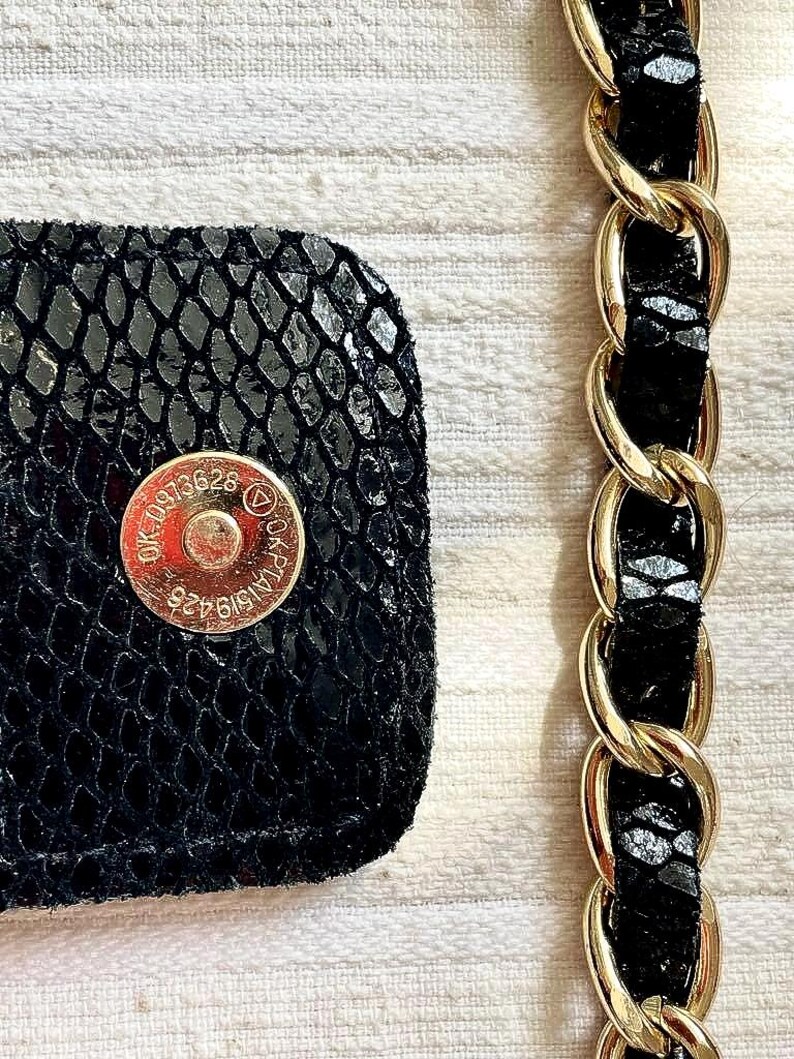 Peut inclure: Gros plan sur un d&eacute;tail de sac &agrave; main noir avec un motif textur&eacute; en peau de serpent. Une bandouli&egrave;re en cha&icirc;ne dor&eacute;e est visible &agrave; droite. Le sac comporte une fermeture magn&eacute;tique dor&eacute;e avec du texte et des chiffres estamp&eacute;s.