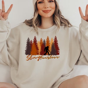 May include: Beige sweatshirt featuring a Bigfoot silhouette walking in front of a forest of brown, orange, and yellow trees. The text "Stay Fearless" is written in an orange script font below the trees. The sweatshirt is a neutral color.