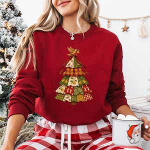 May include: A red sweatshirt featuring a patchwork Christmas tree design with a bird on top. The tree incorporates various patterns and colors, including green, yellow, and red. The person is wearing red and white plaid pajama pants and holding a mug with a Santa Claus design.