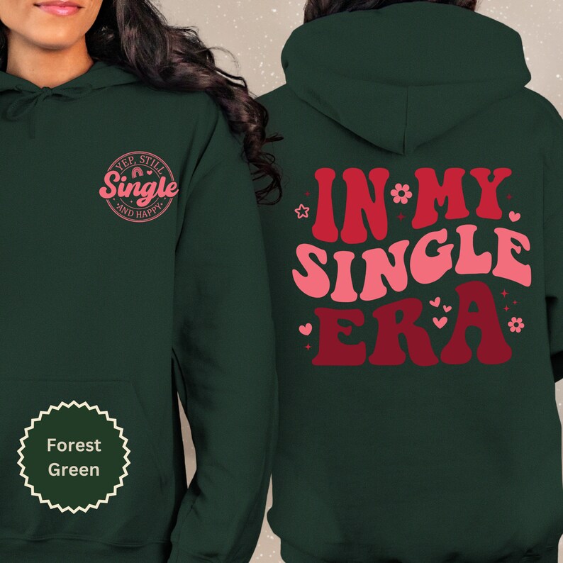 May include: Forest green hoodie with the text "Yep, still single and happy" in a pink circle on the left side and the text "In my single era" in pink and red on the right side.