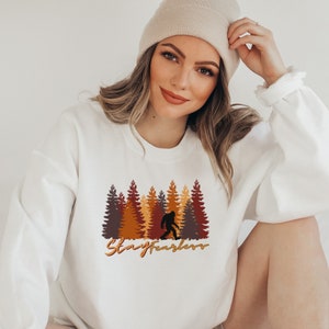 May include: White sweatshirt featuring a Bigfoot graphic in front of a forest of brown, orange, and yellow trees. The text "Stay Fearless" is written in a cursive font below the graphic. The sweatshirt is a crew neck style.