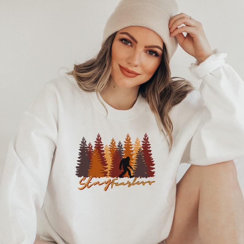 May include: White sweatshirt featuring a Bigfoot graphic in front of a forest of brown, orange, and yellow trees. The text "Stay Fearless" is written in a cursive font below the graphic. The sweatshirt is a crew neck style.