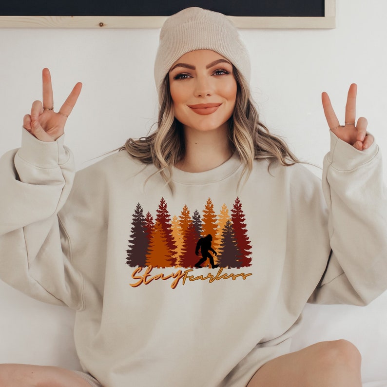 May include: Beige sweatshirt featuring a Bigfoot silhouette walking in front of a forest of brown, orange, and yellow trees. The text "Stay Fearless" is written in an orange script font below the trees. The sweatshirt is a neutral color.