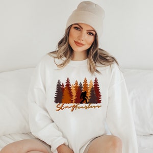 May include: White sweatshirt featuring a Bigfoot silhouette walking in front of a forest of brown, orange, and yellow trees. The text "Stay Fearless" is written in a gold color below the trees.