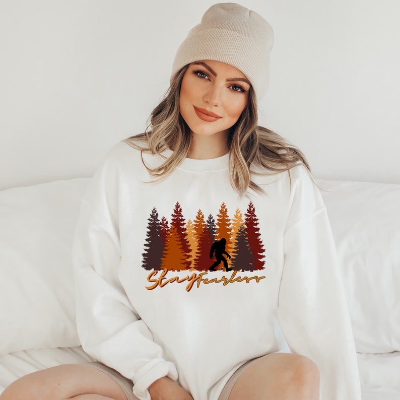 May include: White sweatshirt featuring a Bigfoot silhouette walking in front of a forest of brown, orange, and yellow trees. The text "Stay Fearless" is written in a gold color below the trees.