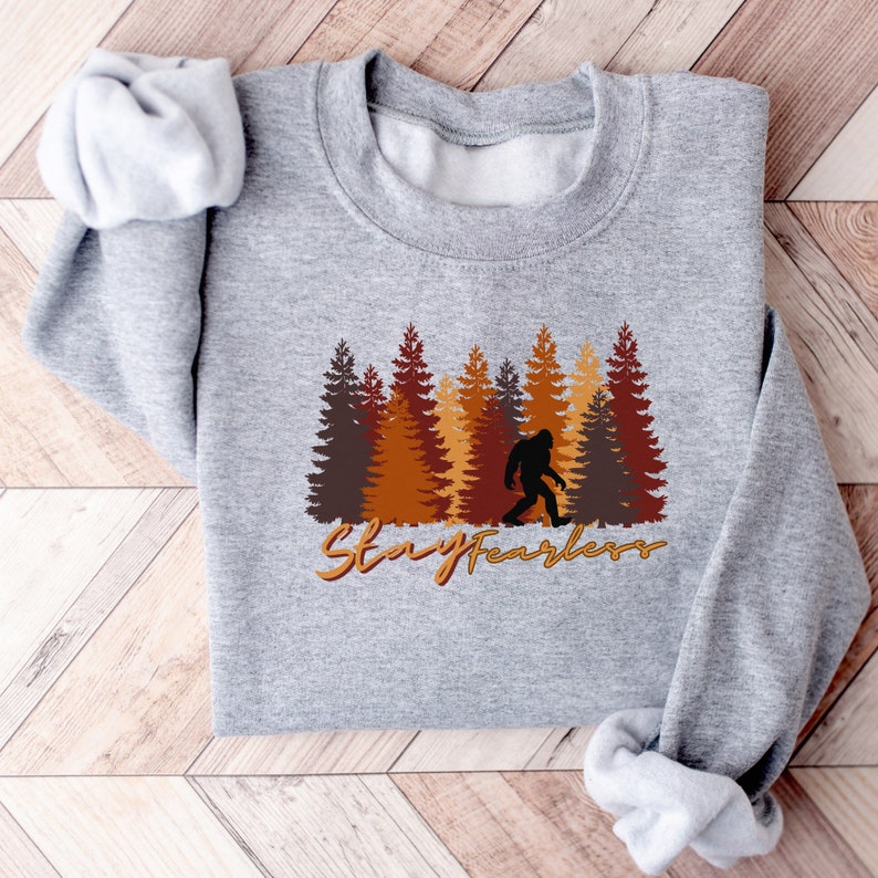 May include: A light gray sweatshirt featuring a forest scene with brown and orange trees and a silhouette of Bigfoot. The words "Stay Fearless" are written in a gold script font across the bottom of the design.