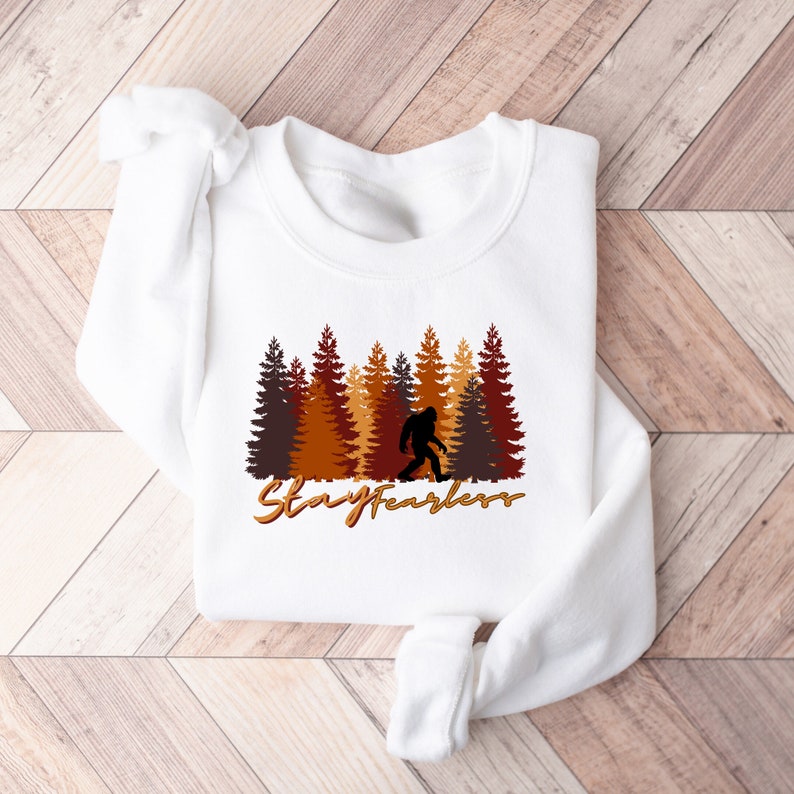 May include: White sweatshirt featuring a forest scene with brown, orange, and yellow trees, a silhouette of Bigfoot, and the text "Stay Fearless" in a gold script. The design is centered on the chest.