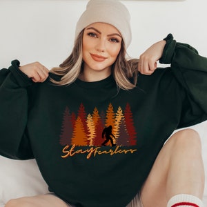 May include: Dark green sweatshirt featuring a Bigfoot silhouette walking in front of a forest of trees in shades of brown and orange. The text "Stay Fearless" is written in a cursive font below the trees.