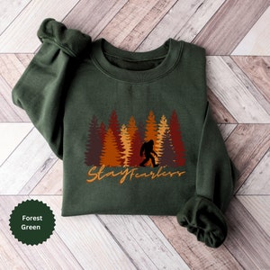 May include: Forest green sweatshirt featuring a Bigfoot silhouette walking in front of a forest of brown, orange, and red trees. The words "Stay Fearless" are written in an orange script font. The sweatshirt is folded on a wooden surface.