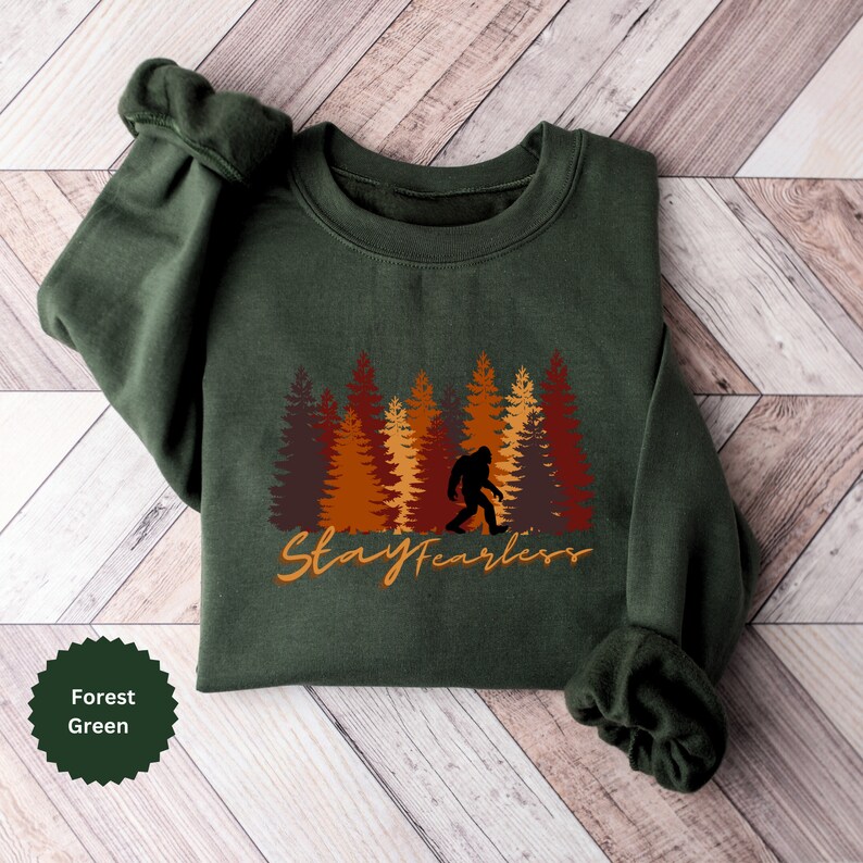 May include: Forest green sweatshirt featuring a Bigfoot silhouette walking in front of a forest of brown, orange, and red trees. The words "Stay Fearless" are written in an orange script font. The sweatshirt is folded on a wooden surface.