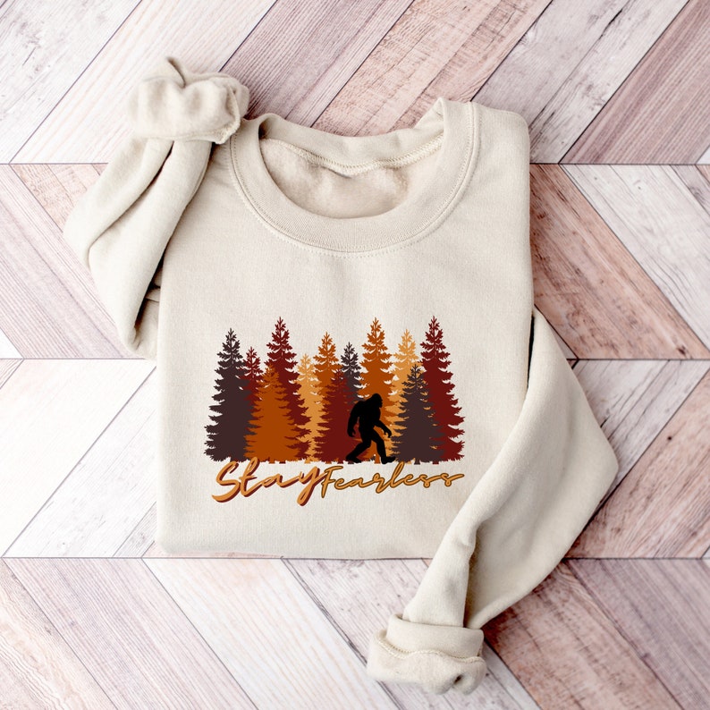 May include: Beige sweatshirt featuring a Bigfoot silhouette walking in front of a forest of brown and orange trees. The text "Stay Fearless" is written in a brown script font below the trees. The sweatshirt is folded on a wooden surface.