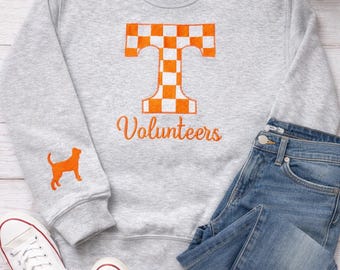 Könnte beinhalten: Graues Sweatshirt mit einem orange-weiß karierten "T" und dem Wort "Volunteers" in orangefarbener Schrift. Auf dem linken Ärmel befindet sich eine orangefarbene Hunde-Silhouette. Das Sweatshirt wird mit blauen Jeans und weißen Turnschuhen kombiniert.