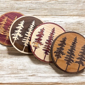 May include: Four round wooden coasters with a laser-cut design of three pine trees. The coasters are made of different types of wood, including cedar, walnut, and maple. The trees are dark brown against the light wood background.