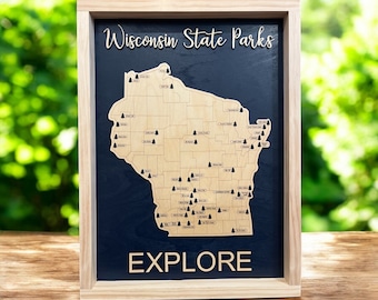 Op de afbeelding: Een houten ingelijste kaart van Wisconsin State Parks met de tekst "Wisconsin State Parks" en "EXPLORE" op een donkerblauwe achtergrond. De kaart is gemaakt van hout en heeft kleine zwarte bomen die de locatie van elk park markeren.