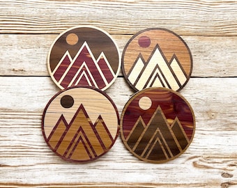 May include: Four round wooden coasters with a mountain and sun design. Each coaster features a different wood grain pattern and color combination. The coasters are approximately 4 inches in diameter.