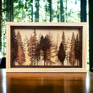 May include: A wooden wall art piece featuring a layered forest scene with multiple trees of varying heights and shades of brown, beige, and tan. The trees are cut out of wood and mounted on a light wood background. The piece is framed in a dark wood frame.