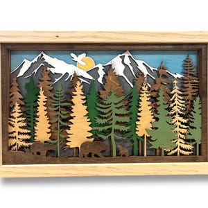May include: A layered wooden art piece depicting a mountain scene with trees, bears, and a sun. The artwork features a light wood frame, with the mountains in white and the sky in blue. The trees are in shades of green and brown.
