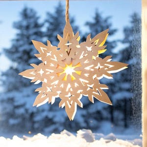 May include: A decorative, wooden snowflake ornament with intricate cut-out designs, suspended by a natural rope. The snowflake is illuminated from within, casting a warm glow. The background shows a snowy winter scene.