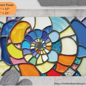 May include: A rectangular memory foam bath mat with a stained glass design. The mat features a spiral pattern in shades of blue, yellow, orange, and red, with dimensions of 24" x 17" and 34" x 21". The design is framed by dark gray lines.