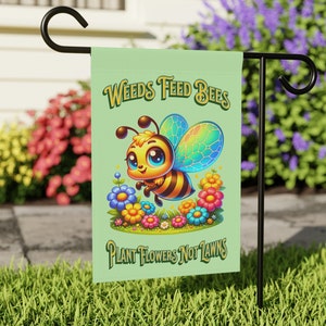 Op de afbeelding: Een groene tuin vlag met een cartoon bij en bloemen. De vlag staat "Weeds Feed Bees Plant Flowers Not Lawns".