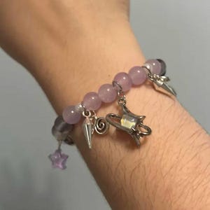 Pu&ograve; includere: Un braccialetto di perline viola con ciondoli argentati, tra cui una stella, una spirale, un cono e un ciondolo pi&ugrave; grande con una stella e una mezzaluna.