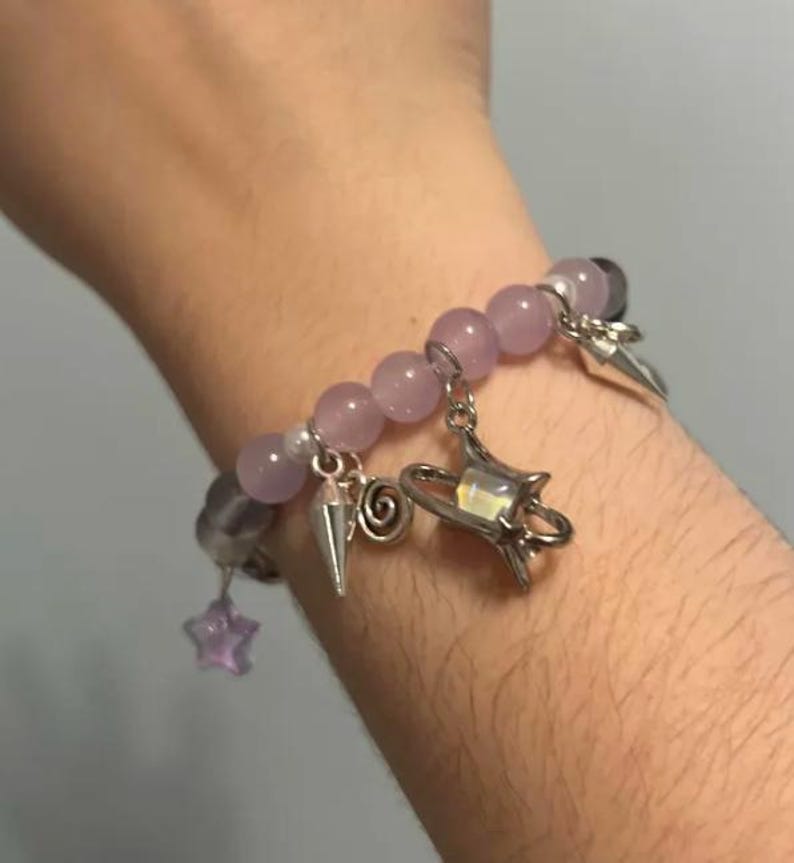 Pu&ograve; includere: Un braccialetto di perline viola con ciondoli argentati, tra cui una stella, una spirale, un cono e un ciondolo pi&ugrave; grande con una stella e una mezzaluna.