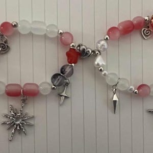 Pu&ograve; includere: Due braccialetti con perline bianche e rosa, ciondoli argentati e un ciondolo a forma di stella rossa. Un braccialetto ha un ciondolo a forma di fiore argentato e l'altro ha un ciondolo a forma di fiore rosa.