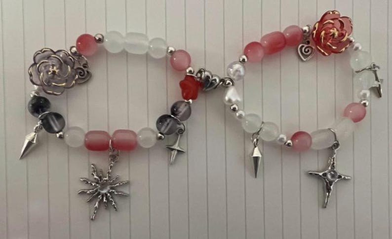 Pu&ograve; includere: Due braccialetti con perline bianche e rosa, ciondoli argentati e un ciondolo a forma di stella rossa. Un braccialetto ha un ciondolo a forma di fiore argentato e l'altro ha un ciondolo a forma di fiore rosa.