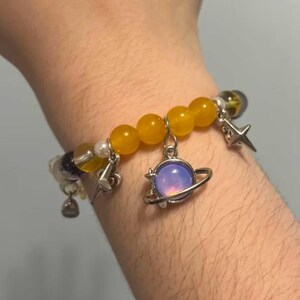 Pu&ograve; includere: Un braccialetto con perline gialle, una perlina viola e bianca e ciondoli argentati. I ciondoli includono una stella, un fulmine e un pianeta con un globo iridescente viola e rosa.