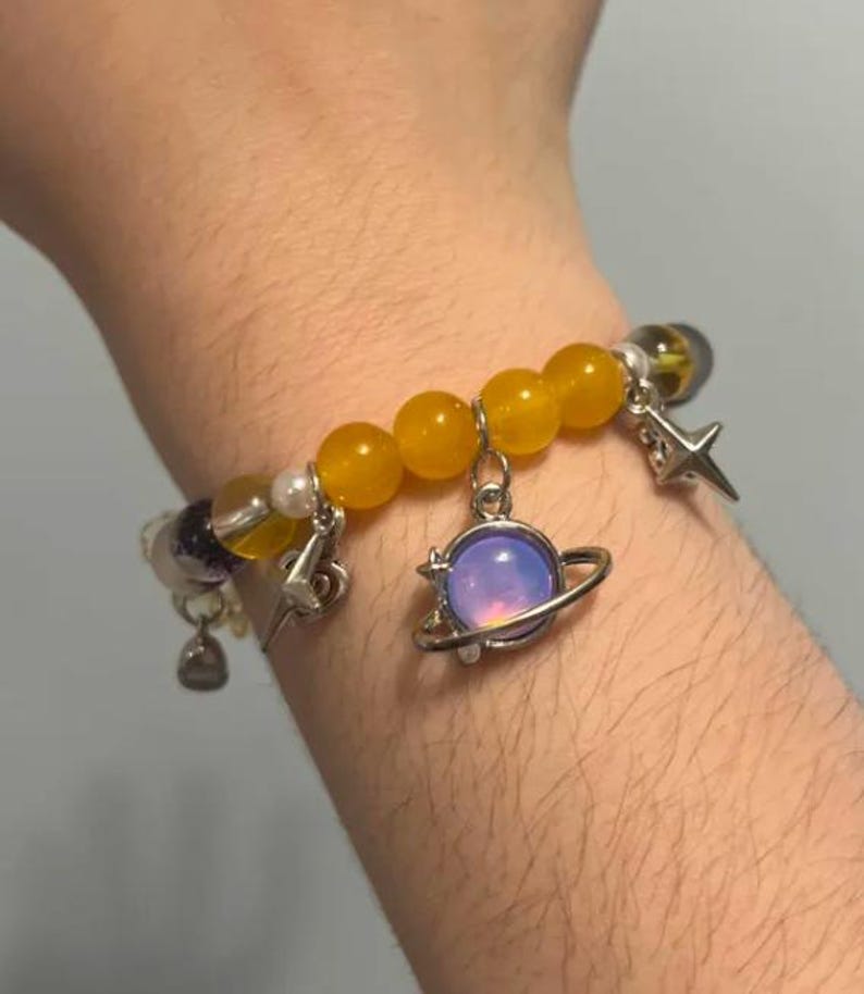 Pu&ograve; includere: Un braccialetto con perline gialle, una perlina viola e bianca e ciondoli argentati. I ciondoli includono una stella, un fulmine e un pianeta con un globo iridescente viola e rosa.