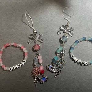 Può includere: Un assortimento di gioielli di perline fatti a mano, tra cui due ciondoli per telefono e due braccialetti. I ciondoli presentano una varietà di perline nei colori rosa, blu e trasparenti, con ciondoli argentati. I braccialetti scrivono "YUKI" e "HITSU".