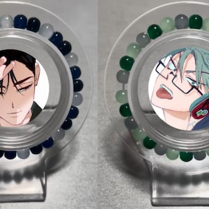 Puede incluir: Dos pulseras con cuentas de diferentes colores e imágenes de personajes de anime. La pulsera de la izquierda tiene cuentas azules y grises y un personaje de anime de cabello negro con una expresión seria. La pulsera de la derecha tiene cuentas verdes y grises y un personaje de anime de cabello verde con una expresión de sorpresa.