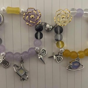 Pu&ograve; includere: Due braccialetti con perline viola e gialle, ciondoli e fiori. Un braccialetto ha un ciondolo a forma di fiore viola e l'altro ha un ciondolo a forma di fiore giallo.