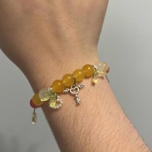 Pu&ograve; includere: Un braccialetto di perline gialle e bianche con un ciondolo a forma di chiave d'argento e un cuore. Il braccialetto ha piccoli ciondoli verdi e bianchi.