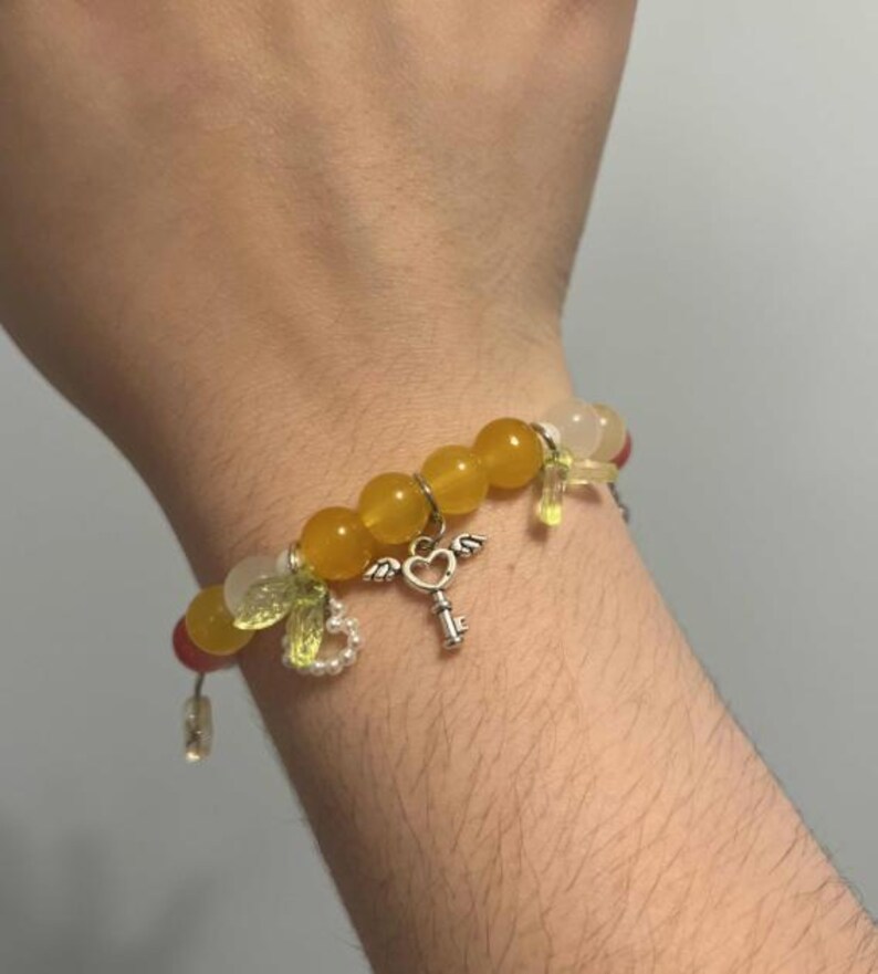 Pu&ograve; includere: Un braccialetto di perline gialle e bianche con un ciondolo a forma di chiave d'argento e un cuore. Il braccialetto ha piccoli ciondoli verdi e bianchi.